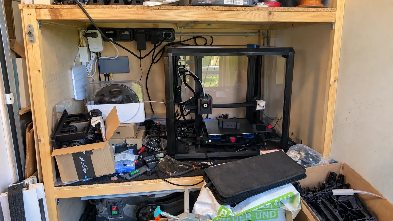 A real home 3D-printing corner with an active printer, boxes of printed parts, cables and the natural clutter of an RC-building hobby in progress.